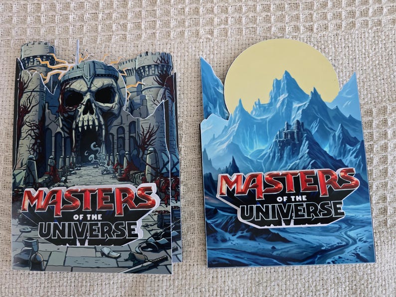 Puede incluir: Dos tarjetas decorativas con el logo "Masters of the Universe". Una tarjeta representa una entrada de castillo en forma de calavera, mientras que la otra muestra una cordillera con un castillo y una luna amarilla. El logo es rojo y blanco.