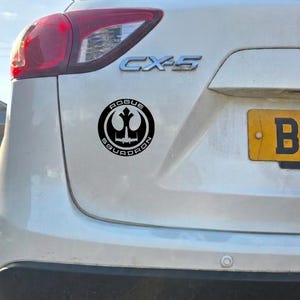 May include: A circular black and white vinyl decal applied to the rear of a white vehicle. The sticker features a stylized emblem with a central starfighter silhouette and the text Rogue Squadron arranged in a circular border around the design.