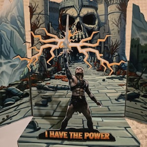 Puede incluir: Tarjeta desplegable con una figura musculosa sosteniendo una espada, con un fondo de castillo y el texto "I HAVE THE POWER". La tarjeta tiene una combinaci&oacute;n de colores gris y naranja.