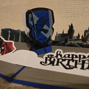 May include: A pop-up birthday card featuring a blue and black crest with a bird emblem, a figure in a red cloak, and a castle silhouette. The words "happy BIRTHDAY" are cut out in black script.