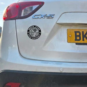 May include: A circular black and white vinyl decal applied to the rear of a white vehicle. The sticker features a graphic of the Death Star with the text "DEATH STAR" arched across the top and "TECH SUPPORT" arched across the bottom.
