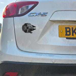 May include: A white Mazda CX-5 vehicle features a black decal on the rear hatch. The decal is shaped like a spaceship and contains the text MY OTHER RIDE IS THE FALCON. The car has a red taillight and a yellow license plate partially visible.