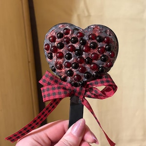 Puede incluir: Un objeto decorativo en forma de corazón con un tallo negro y un lazo a cuadros rojo y negro. El corazón está cubierto de cuentas y purpurina rojas y negras, creando una superficie texturizada.
