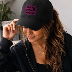 May include: A black baseball cap featuring a pink embroidered rectangular frame with scalloped edges and the text "SOMEBODY'S PROBLEM" in a bold, sans-serif font centred within the design on the front panel of the hat.