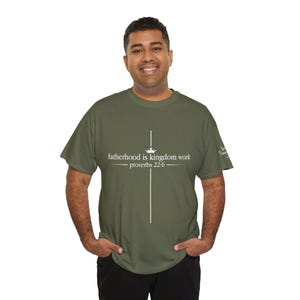 Puede incluir: Una camiseta de manga corta color verde oliva con un gráfico blanco de una cruz con un pequeño icono de corona sobre el texto "fatherhood is kingdom work" y "proverbs 22:6" centrado en el pecho, con texto blanco adicional en la manga.