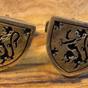 May include: A pair of gold-toned metal cufflinks, each shaped like a shield. The surface of each cufflink features a recessed black enamel design depicting a rampant lion, with a smooth metallic border framing the shield-shaped emblem.