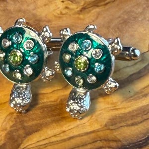 May include: A pair of silver cufflinks with a turtle design. The turtles have a green enamel shell with clear and green gemstones. The cufflinks are set against a wooden background.