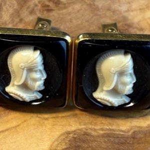 May include: A pair of square cufflinks with a black background and gold trim. Each cufflink features a white cameo of a Roman soldier's profile. The cufflinks are set against a wooden surface.