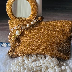 May include: A gold beaded clutch bag featuring a circular handle with an integrated mirror. The bag is adorned with a gold-toned chain strap accented with white faux pearls and a gold script charm that reads Givenchy. A strand of white pearls rests below.