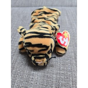 May include: A plush tiger toy with orange and black stripes, featuring small ears and a black nose. A red heart-shaped tag with the word "Ty" is attached to the toy. The toy is lying on a gray surface.
