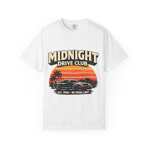 May include: White t-shirt with a graphic featuring a black sports car, palm trees, and a sunset design. The text "MIDNIGHT DRIVE CLUB" is above the car, with "EST. 1998 - NO SPEED LIMIT" below. The shirt has a crew neck.