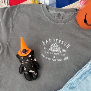 May include: A charcoal gray t-shirt featuring a white graphic that reads Sanderson Witch Museum est. 1693 Home of the black flame candle. A small black plush cat doll wearing a bright orange witch hat with a purple band is placed on the shirt.