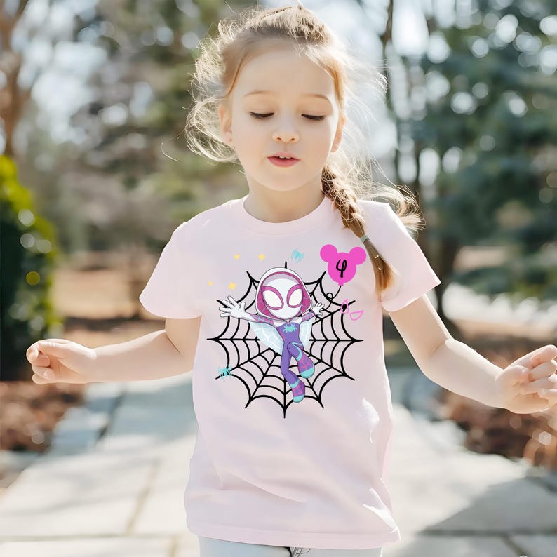 Op de afbeelding: Lichtroze t-shirt met een cartoon superheldenafbeelding. Het ontwerp toont een personage in een spinnenweb, met een wit masker en een paars en blauw pak. Een roze ballon met het nummer 4 is ook zichtbaar.