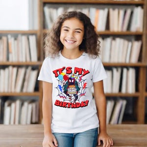 K&ouml;nnte beinhalten: Wei&szlig;es T-Shirt mit einer farbenfrohen Grafik, die einen Superhelden mit einem Geburtstagskuchen zeigt, umgeben von einem Spinnennetz-Design. Der Text "IT'S MY BIRTHDAY" ist rot dargestellt. Das Shirt hat kurze &Auml;rmel.