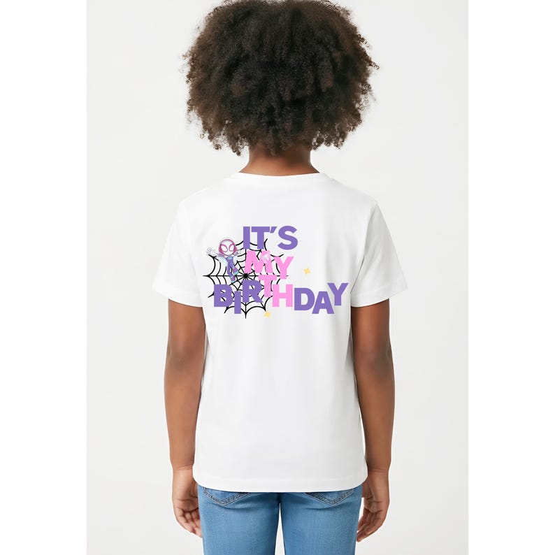 Op de afbeelding: Wit T-shirt met de tekst "IT'S MY BIRTHDAY" in paars en roze, met een spinnenwebontwerp. Het shirt heeft een stripfiguur. De persoon draagt een blauwe spijkerbroek.