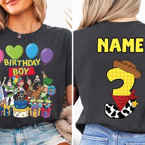 Op de afbeelding: Donkergrijs T-shirt met een kleurrijke afbeelding met ballonnen, de tekst "BIRTHDAY BOY" en stripfiguren. De achterkant van de shirt heeft het woord "NAME" en een nummer 3 met een cowboyhoed en bandana.