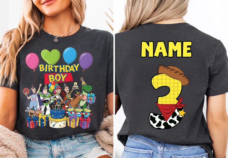 Op de afbeelding: Donkergrijs T-shirt met een kleurrijke afbeelding met ballonnen, de tekst "BIRTHDAY BOY" en stripfiguren. De achterkant van de shirt heeft het woord "NAME" en een nummer 3 met een cowboyhoed en bandana.
