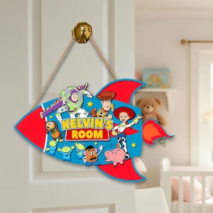 May include: A colorful wooden sign in the shape of a rocket, featuring characters from the Toy Story movie. The sign is red and blue, with the text "KELVIN'S ROOM". It hangs from a gold-colored metal hook.