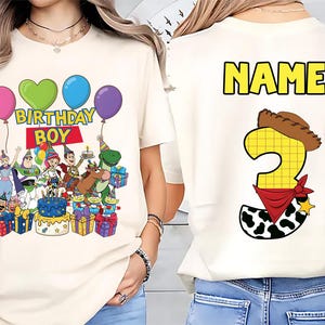 Op de afbeelding: Cr&egrave;mekleurig T-shirt met een kleurrijke afbeelding van Toy Story-personages, ballonnen en de tekst "BIRTHDAY BOY". De achterkant toont een groot nummer 3 met een cowboyhoed en bandana, en het woord "NAME" in geel.
