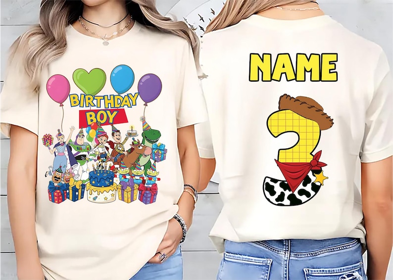 Op de afbeelding: Cr&egrave;mekleurig T-shirt met een kleurrijke afbeelding van Toy Story-personages, ballonnen en de tekst "BIRTHDAY BOY". De achterkant toont een groot nummer 3 met een cowboyhoed en bandana, en het woord "NAME" in geel.