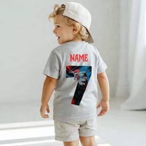 K&ouml;nnte beinhalten: Ein graues T-Shirt mit einer Grafik eines Superhelden und der Zahl 7. Das Wort "NAME" ist rot &uuml;ber der Grafik aufgedruckt. Das Shirt wird von einem kleinen Kind getragen, das eine wei&szlig;e Baseballkappe und khakifarbene Shorts tr&auml;gt.