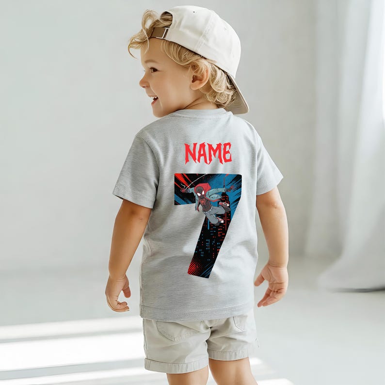 K&ouml;nnte beinhalten: Ein graues T-Shirt mit einer Grafik eines Superhelden und der Zahl 7. Das Wort "NAME" ist rot &uuml;ber der Grafik aufgedruckt. Das Shirt wird von einem kleinen Kind getragen, das eine wei&szlig;e Baseballkappe und khakifarbene Shorts tr&auml;gt.