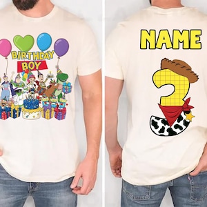 Op de afbeelding: Cr&egrave;mekleurig T-shirt met een kleurrijke afbeelding van Toy Story-personages, ballonnen en de tekst "BIRTHDAY BOY" op de voorkant. De achterkant van het shirt toont het woord "NAME" en het cijfer 3 met een cowboyhoed en bandana.