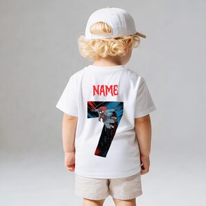K&ouml;nnte beinhalten: Wei&szlig;es T-Shirt mit einer Grafik eines Superhelden und der Zahl 7. Das Wort "NAME" ist rot &uuml;ber der Grafik aufgedruckt. Das Kind tr&auml;gt eine wei&szlig;e Baseballkappe und beige Shorts.