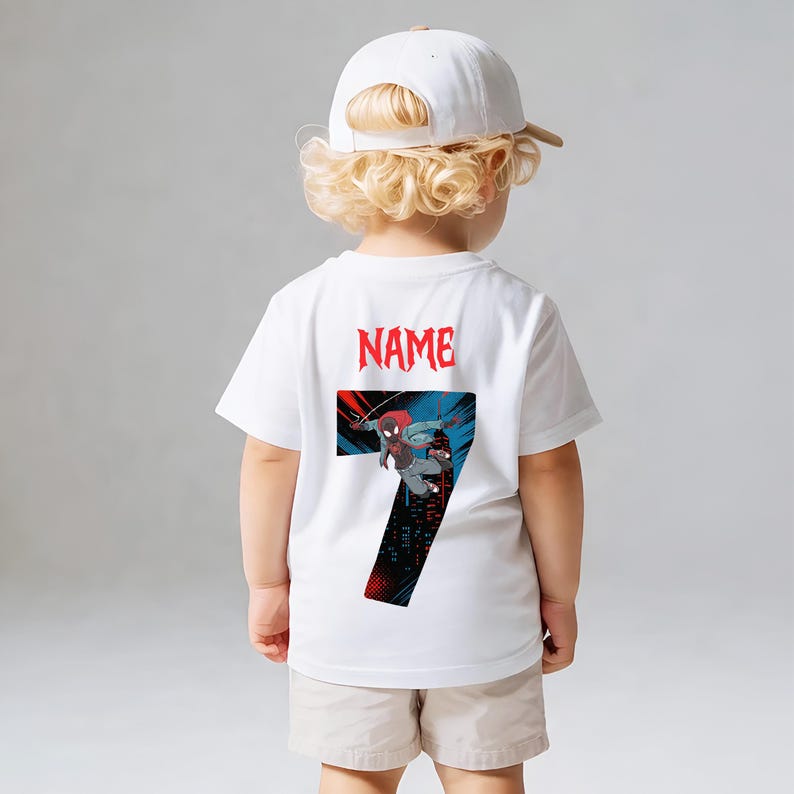 K&ouml;nnte beinhalten: Wei&szlig;es T-Shirt mit einer Grafik eines Superhelden und der Zahl 7. Das Wort "NAME" ist rot &uuml;ber der Grafik aufgedruckt. Das Kind tr&auml;gt eine wei&szlig;e Baseballkappe und beige Shorts.