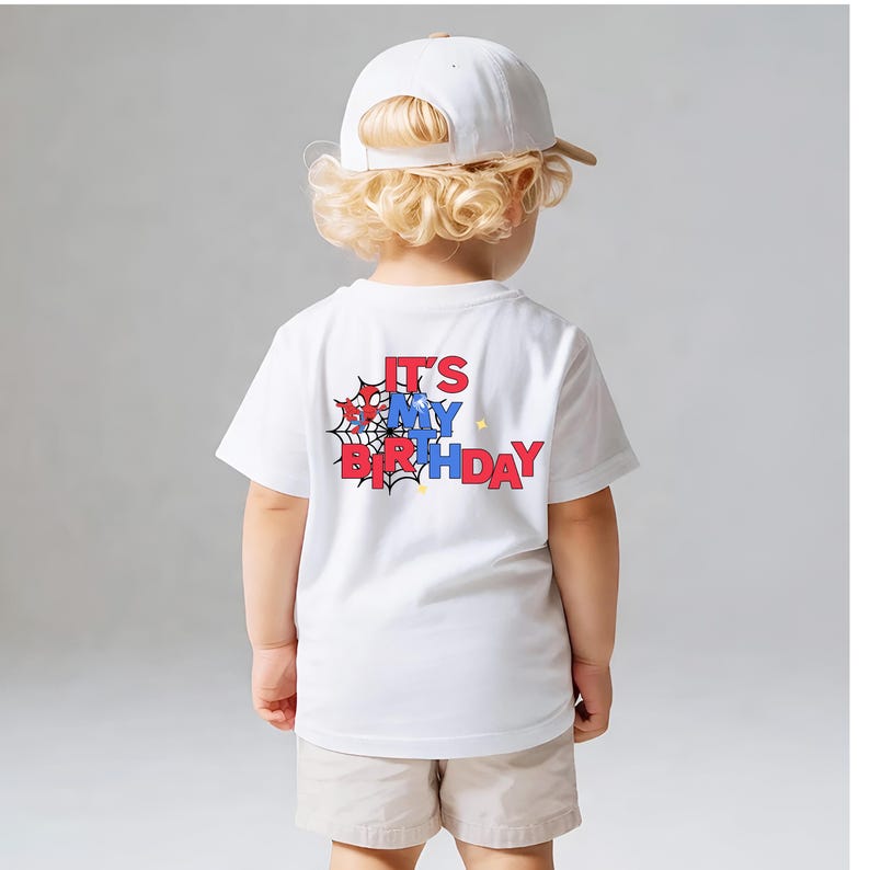 Op de afbeelding: Een wit T-shirt met de tekst "IT'S MY BIRTHDAY" in rood en blauw, met een spinnenwebontwerp. Het shirt wordt gedragen door een kind dat een witte baseballpet en beige shorts draagt.