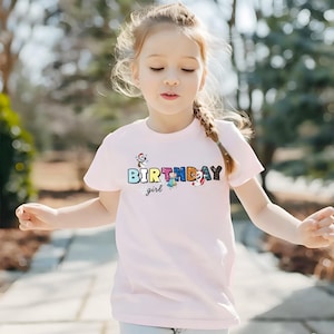 Op de afbeelding: Een lichtroze t-shirt met het woord "BIRTHDAY" in kleurrijke letters en het woord "girl" in cursief. Het shirt heeft cartoonafbeeldingen van een sneeuwpop, een eenhoorn en een rendier. Het meisje draagt het t-shirt.