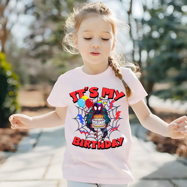 K&ouml;nnte beinhalten: Hellrosa T-Shirt mit einem Superhelden-Motiv, das eine Geburtstagstorte, Luftballons und ein Spinnennetz-Design zeigt. Der Text "IT'S MY BIRTHDAY" ist rot gedruckt.