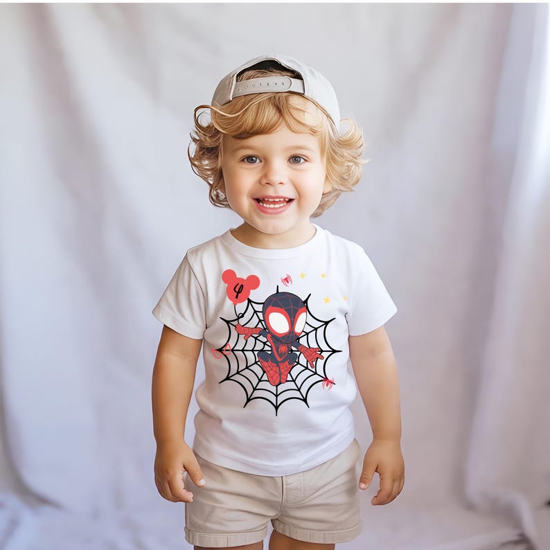 Op de afbeelding: Een lachend kind draagt een wit t-shirt met een Spider-Man-afbeelding op een web, met het nummer "4" op een ballon. Het kind draagt ook een kaki korte broek en een grijze baseball cap.