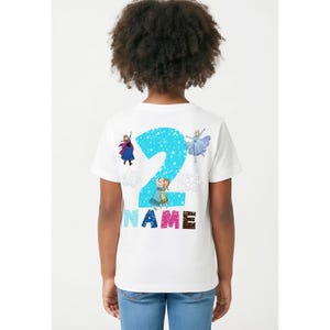 Op de afbeelding: Wit T-shirt met een groot blauw nummer 2, sneeuwvlokkenpatroon en tekenfilmfiguren. Het woord "NAME" staat eronder in kleurrijke letters. Het shirt is ontworpen voor een kind.