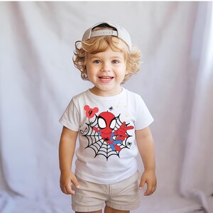 Op de afbeelding: Een lachend kind draagt een wit t-shirt met een Spider-Man-afbeelding. Het shirt heeft een rood-blauw Spider-Man-ontwerp met een spinnenweb. Het kind draagt ook een kaki korte broek en een grijze baseballcap.