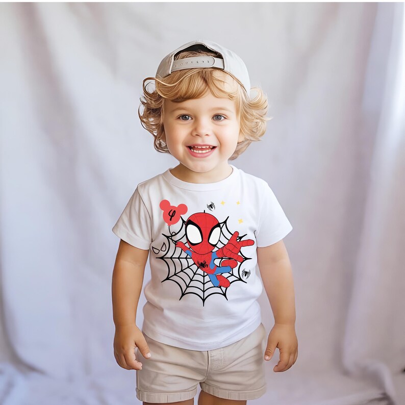 Op de afbeelding: Een lachend kind draagt een wit t-shirt met een Spider-Man-afbeelding. Het shirt heeft een rood-blauw Spider-Man-ontwerp met een spinnenweb. Het kind draagt ook een kaki korte broek en een grijze baseballcap.