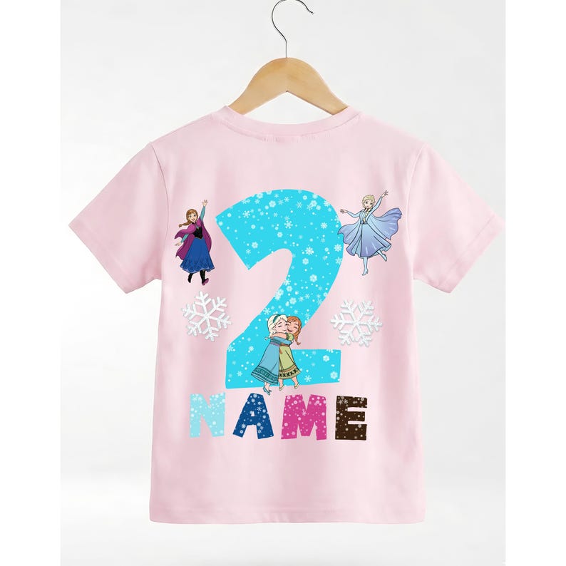 Op de afbeelding: Roze T-shirt met korte mouwen met een groot blauw nummer 2 met een sneeuwvlokontwerp. Het shirt heeft illustraties van personages uit de film Frozen, met het woord "NAME" in kleurrijke blokletters eronder.