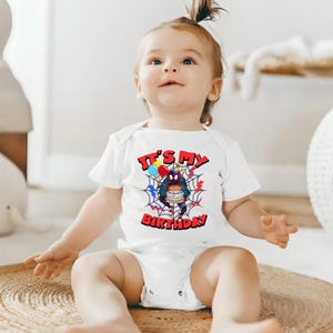 K&ouml;nnte beinhalten: Wei&szlig;er Baby-Body mit dem Text "IT'S MY BIRTHDAY" in Rot. Das Design zeigt eine Cartoon-Spinne, die einen Kuchen h&auml;lt, umgeben von einem Spinnennetz und Luftballons. Der Body hat kurze &Auml;rmel.