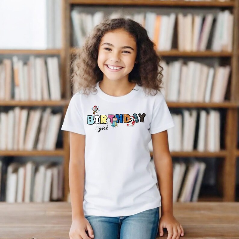 Op de afbeelding: Een wit t-shirt met de woorden "BIRTHDAY girl" in kleurrijke letters en afbeeldingen. Het shirt wordt gedragen door een lachende persoon. De achtergrond is een boekenkast.