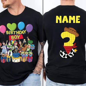 Op de afbeelding: Zwart T-shirt met een kleurrijke afbeelding met personages, ballonnen en de tekst "BIRTHDAY BOY" op de voorkant. De achterkant toont "NAME" boven een nummer 3 met een cowboyhoed en bandana.