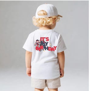 Op de afbeelding: Een wit t-shirt met de tekst "IT'S MY BIRTHDAY" in rood en zwart, met een spinnenwebontwerp. Het shirt wordt gedragen door een kind dat een witte baseballpet en beige shorts draagt.