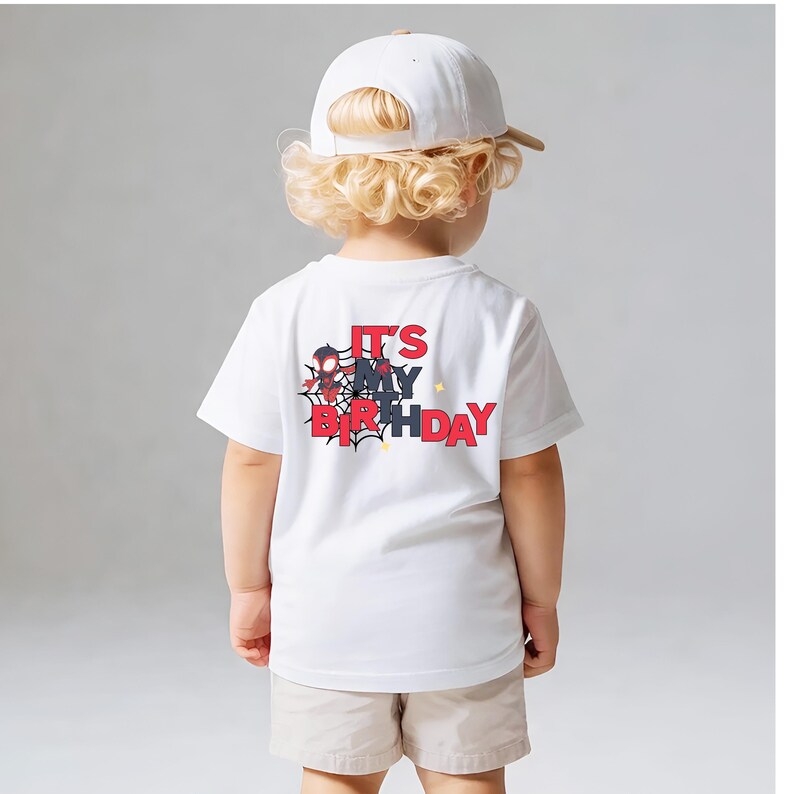 Op de afbeelding: Een wit t-shirt met de tekst "IT'S MY BIRTHDAY" in rood en zwart, met een spinnenwebontwerp. Het shirt wordt gedragen door een kind dat een witte baseballpet en beige shorts draagt.