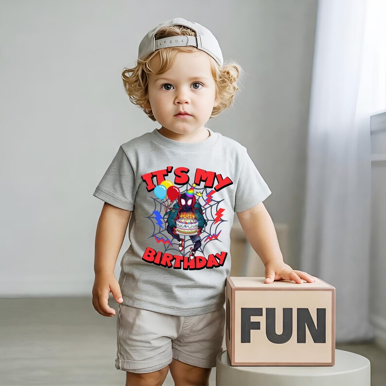 K&ouml;nnte beinhalten: Ein Kind tr&auml;gt ein hellgraues T-Shirt mit dem Text "IT'S MY BIRTHDAY" und einem Spider-Man-Motiv. Das Kind tr&auml;gt auch khakifarbene Shorts und eine graue Baseballkappe. Ein Holzblock mit dem Wort "FUN" befindet sich neben dem Kind.