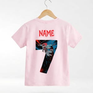 K&ouml;nnte beinhalten: Hellrosa T-Shirt mit einem Superhelden-Motiv, der Zahl 7 und dem Wort "NAME" in Rot. Der Superheld wird in einer dynamischen Pose vor einem Stadtbildhintergrund dargestellt.
