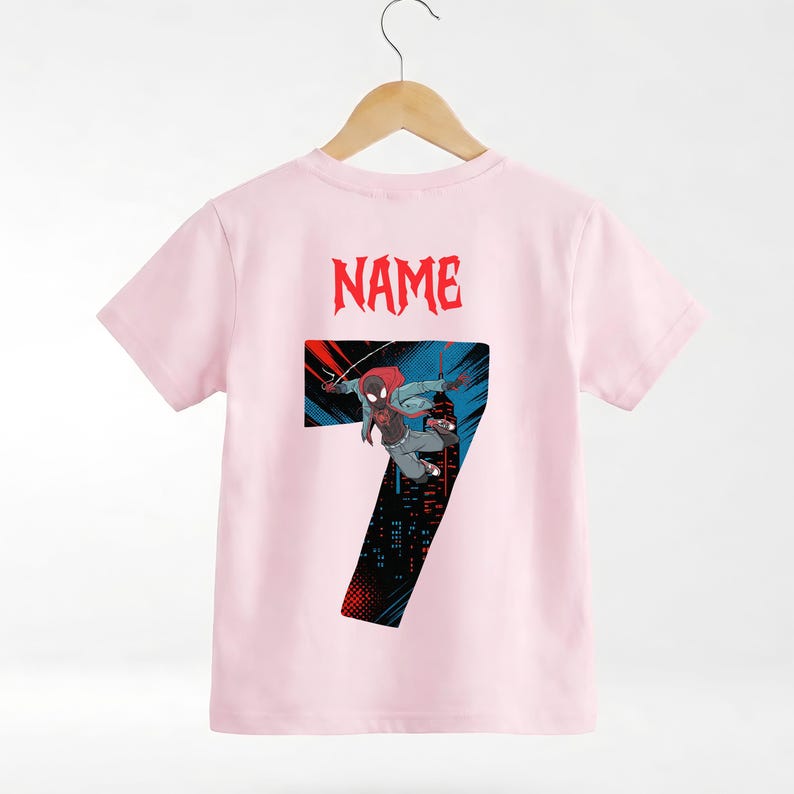 K&ouml;nnte beinhalten: Hellrosa T-Shirt mit einem Superhelden-Motiv, der Zahl 7 und dem Wort "NAME" in Rot. Der Superheld wird in einer dynamischen Pose vor einem Stadtbildhintergrund dargestellt.