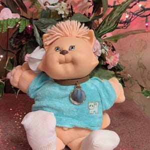 Op de afbeelding: Een pluche kattenpop met perzikkleurige huid en garen haar, gekleed in een turquoise shirt met korte mouwen en een kleine witte kattenafbeelding. De pop heeft een bruine halsband met een ronde zilverkleurige hanger en draagt witte stoffen sokken.