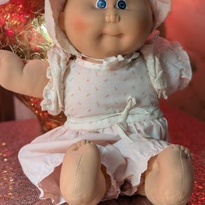 Könnte beinhalten: Eine Stoffpuppe mit rundem Gesicht, blauen Augen und blondem Wollhaar. Die Puppe trägt eine weiße Haube mit Lochstickerei, ein weißes Kleid mit kleinem rosa Blumenmuster sowie ein weißes Bindeband in der Taille und Ärmel mit Rüschen.