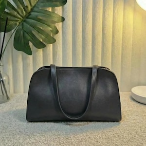 Puede incluir: Un bolso de mano gris oscuro con dos asas. El bolso tiene una forma rectangular redondeada y una textura suave. El fondo incluye una planta verde, una lámpara y una pared texturizada.