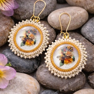Pode incluir: Brincos redondos com um design de abelha e flores. Os brincos têm uma moldura dourada e uma borda com contas creme. O fundo inclui pedras cinzentas e bege e flores roxas.
