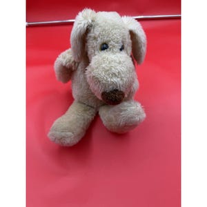 May include: A plush dog toy with cream-colored, textured fur, floppy ears, and a dark brown nose. The stuffed animal features large, dark eyes and a soft, rounded snout, designed with a seated posture and a fuzzy, tactile exterior finish.